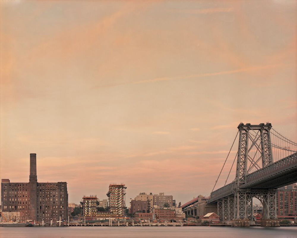 bridge and series of buildings on the waterfront are lit by an orange-purple sunset