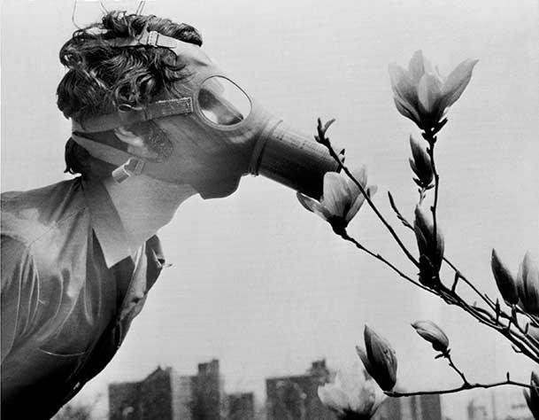 Black and white photograph of a man wearing a gas mask leaning over to smell flowers. In the background is a city skyline.