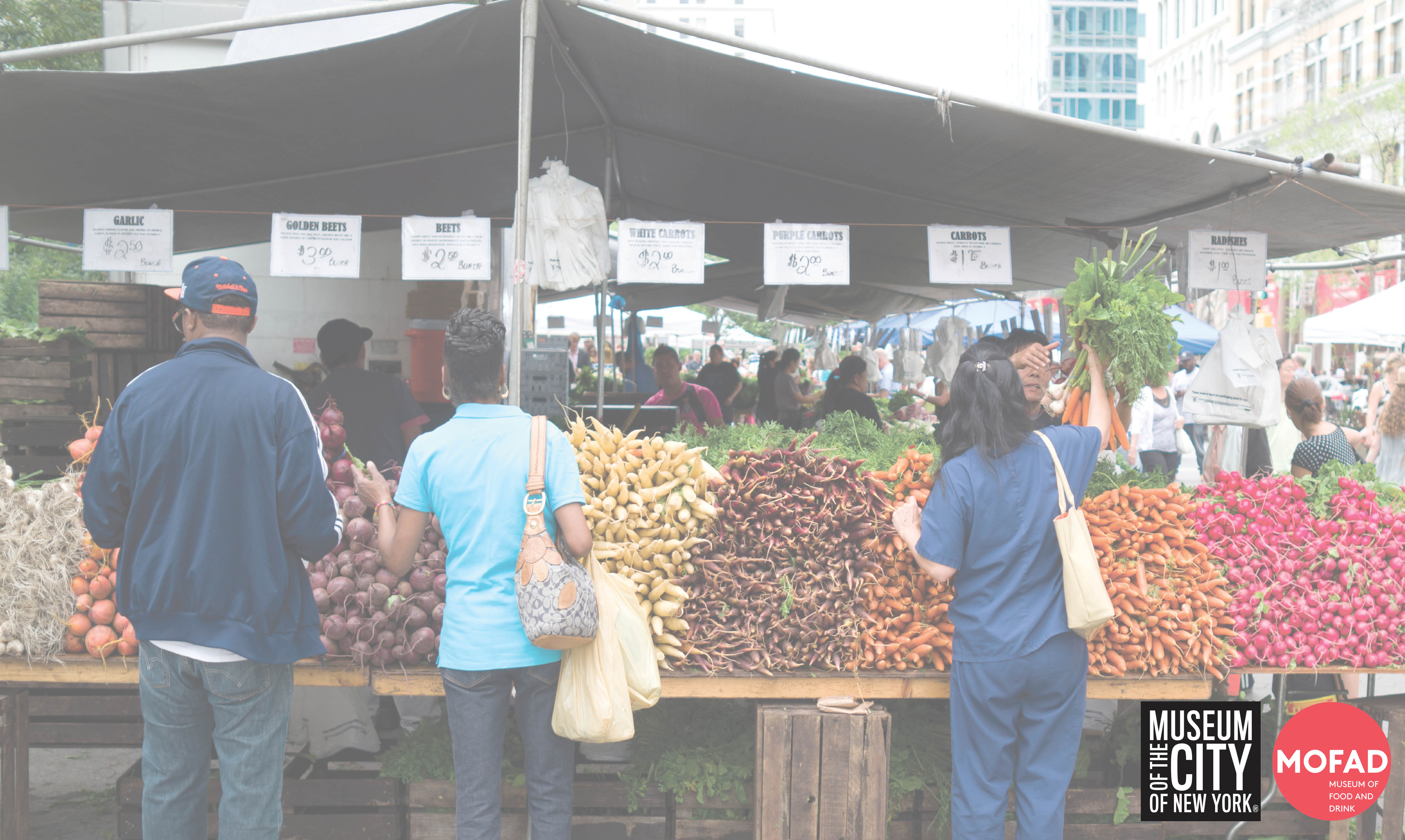With their backs to the camera, three people are standing in front of a farmer’s market booth at the Union Square Greenmarket. There are stacks of garlic, beets, and carrots from left to right.  