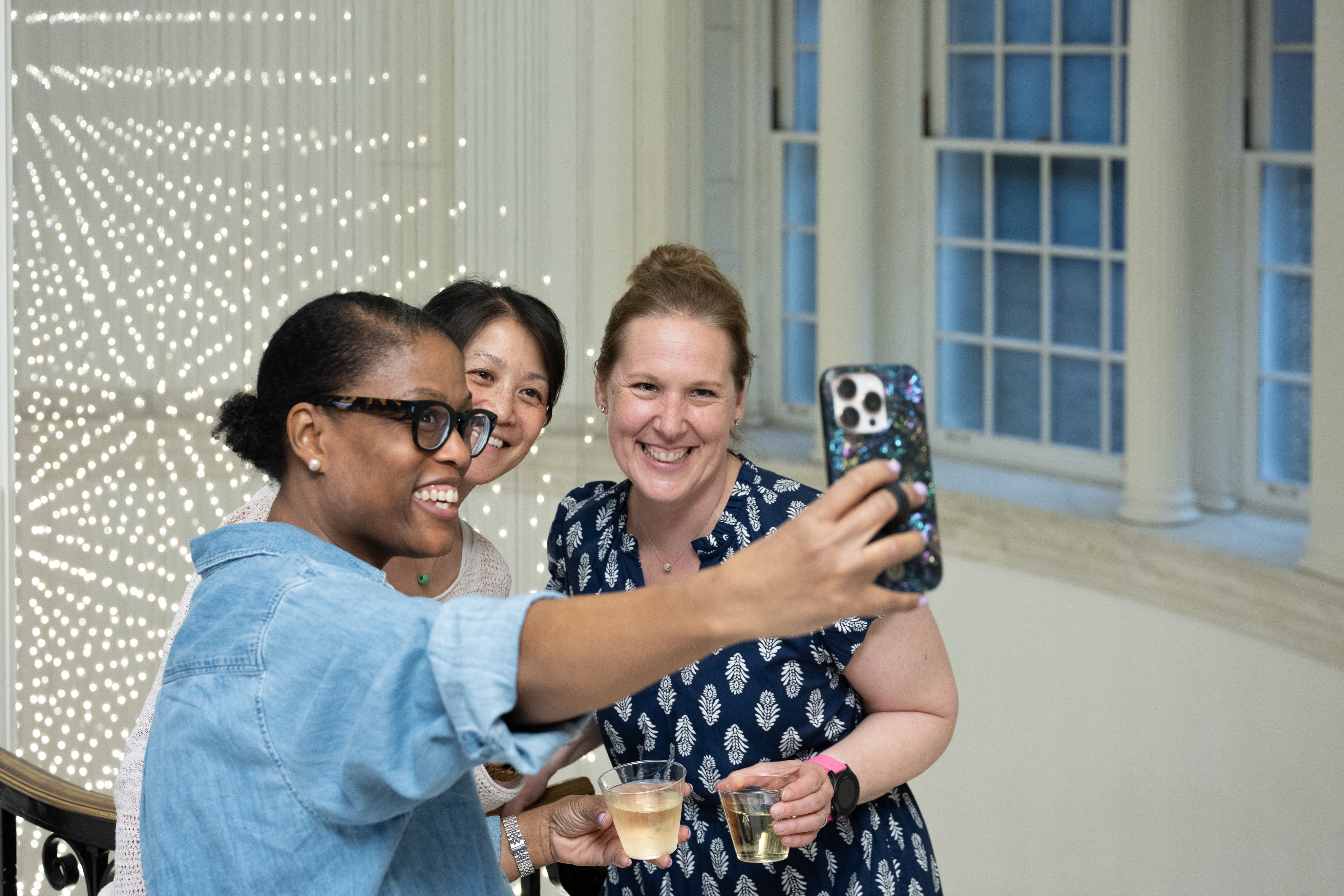 Three attendees smile for a selfie with drinks in the Museum’s Marble Court, with Starlight glowing in the background.