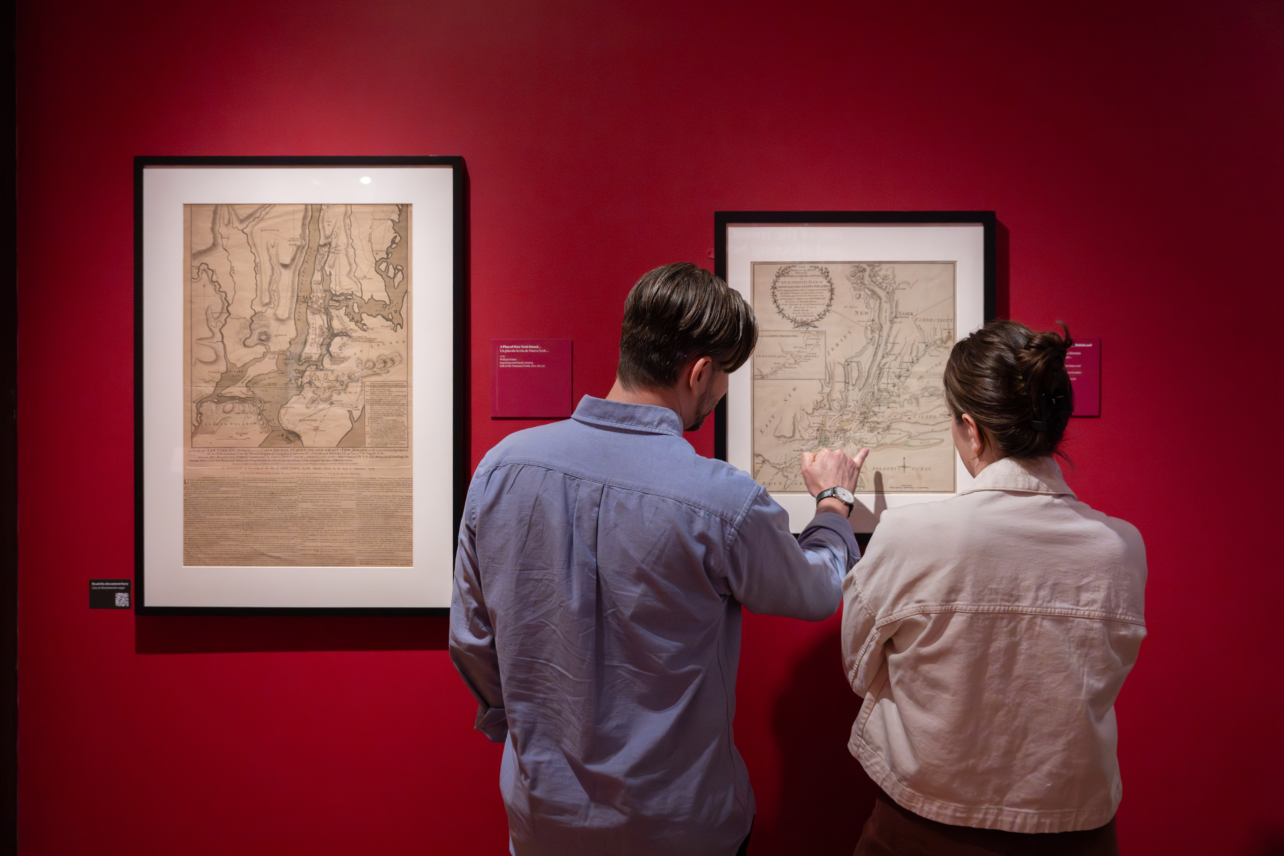 Two teachers view and discuss framed historic maps displayed on a red gallery wall in a museum exhibition.