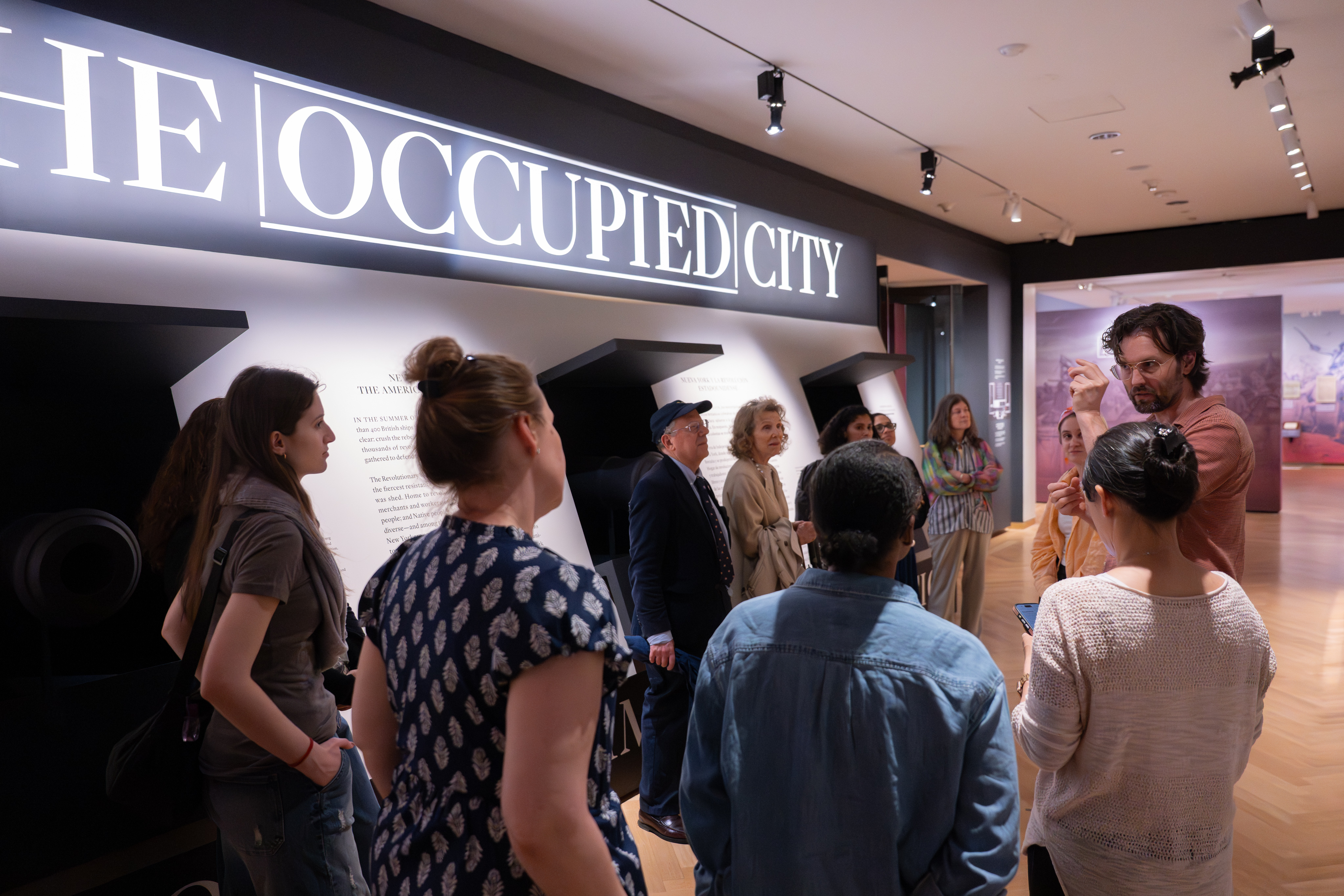 A group of visitors stands in a museum gallery beneath a large illuminated sign reading “THE OCCUPIED CITY,” listening to a curator and viewing wall-mounted exhibition panels in a modern, well-lit space.
