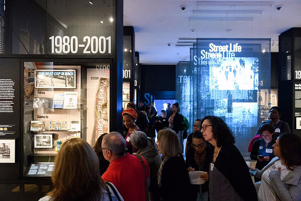 A group of educators gathered in the Museum of the City of New York’s “World City” gallery. Prominent displays include sections titled “1980–2001” and “Street Life,” with exhibits featuring themes such as policing, the events of 9/11, and urban life. The room is dimly lit with illuminated display cases and projected images. Attendees appear engaged, reading, discussing, and taking notes. 
