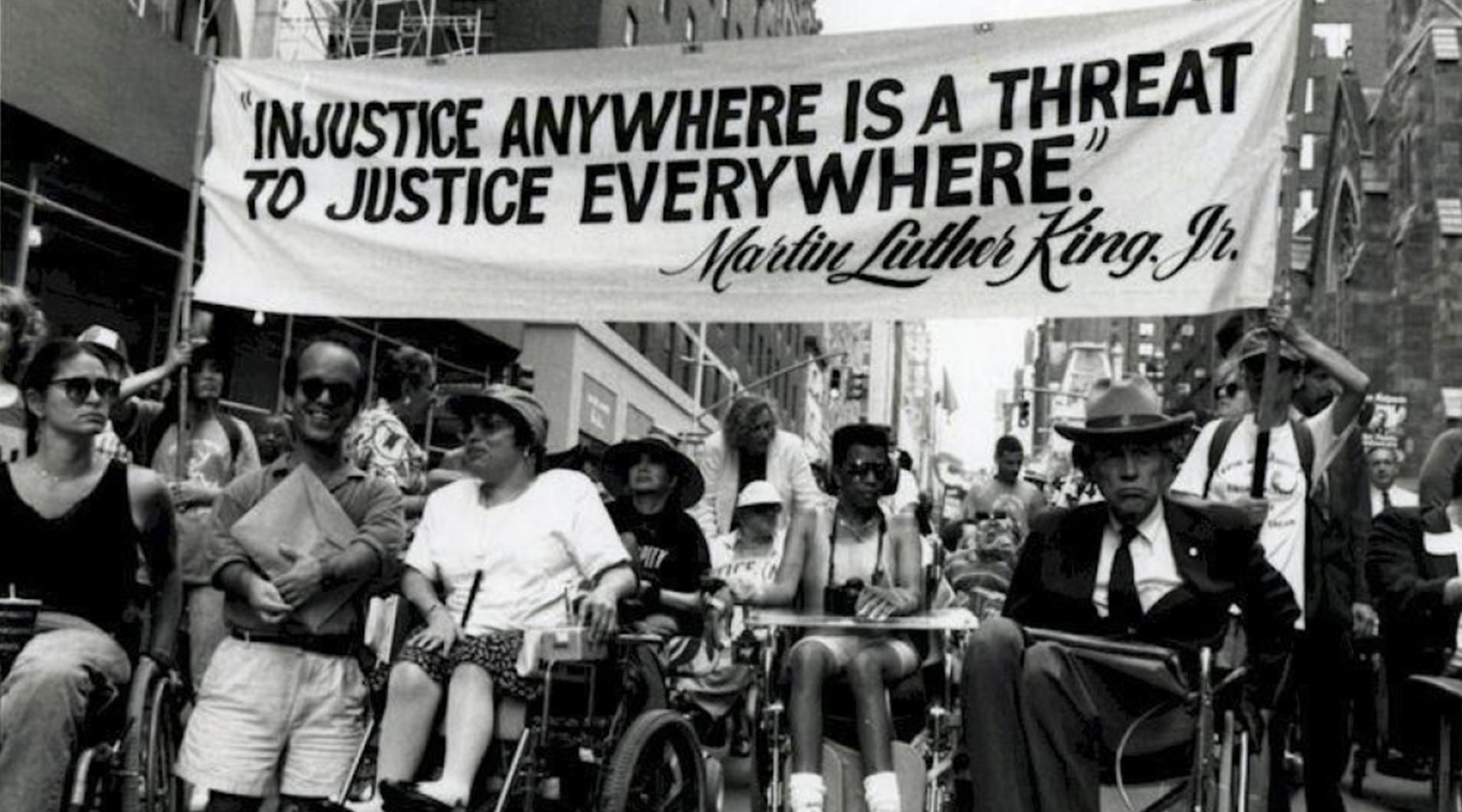 A crowd of people with disabilities and individuals in wheelchairs gather under a banner that reads "Injustice Anywhere is a Threat to Justice Everywhere" Martin Luther King Jr.