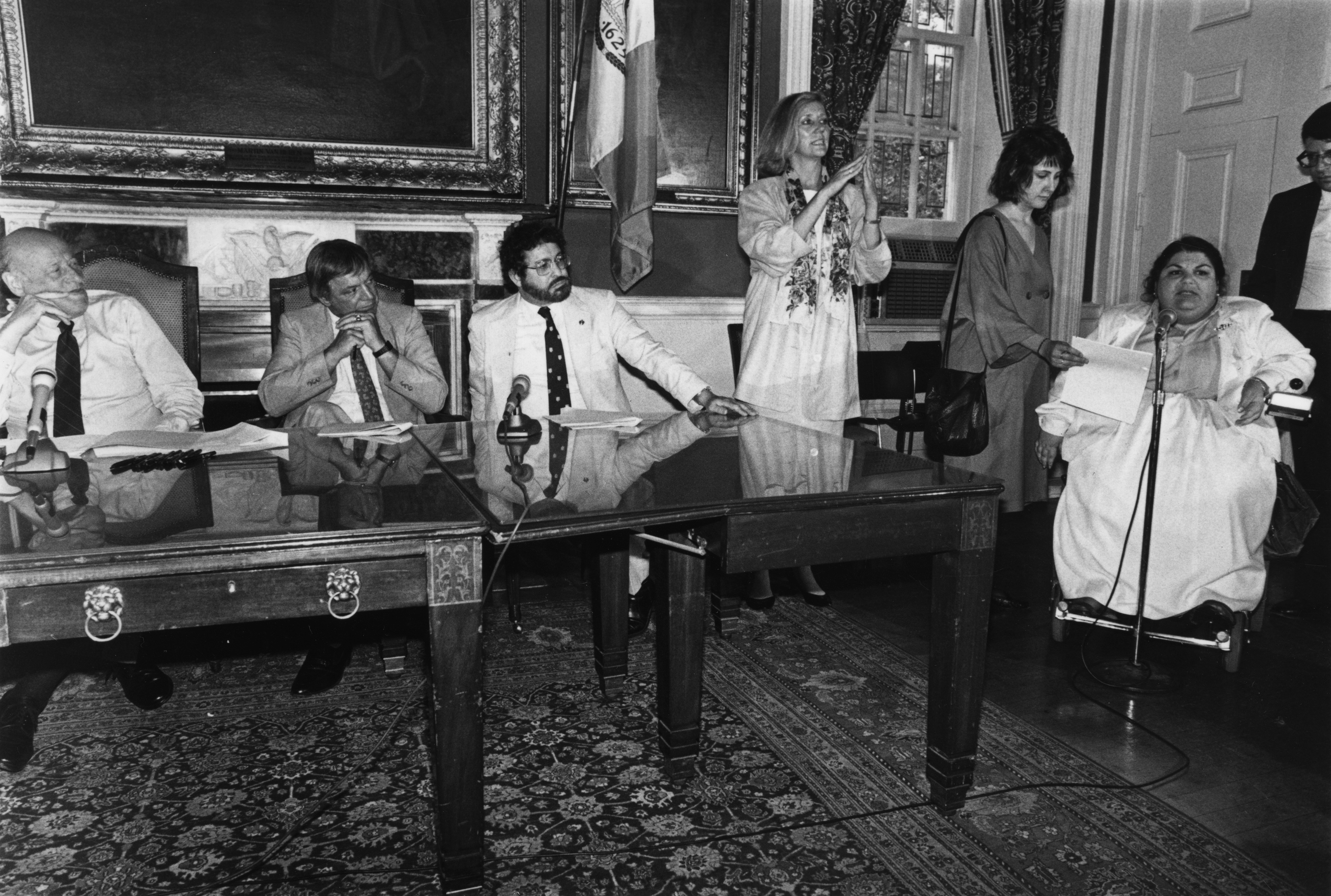 In an ornately decorated room, three men in suits sit at tables to the left with microphones. To there right, a woman stands, signing, while further right, another women holds paper/speech for a third woman in a wheelchair, who speaks into a mic stand. 