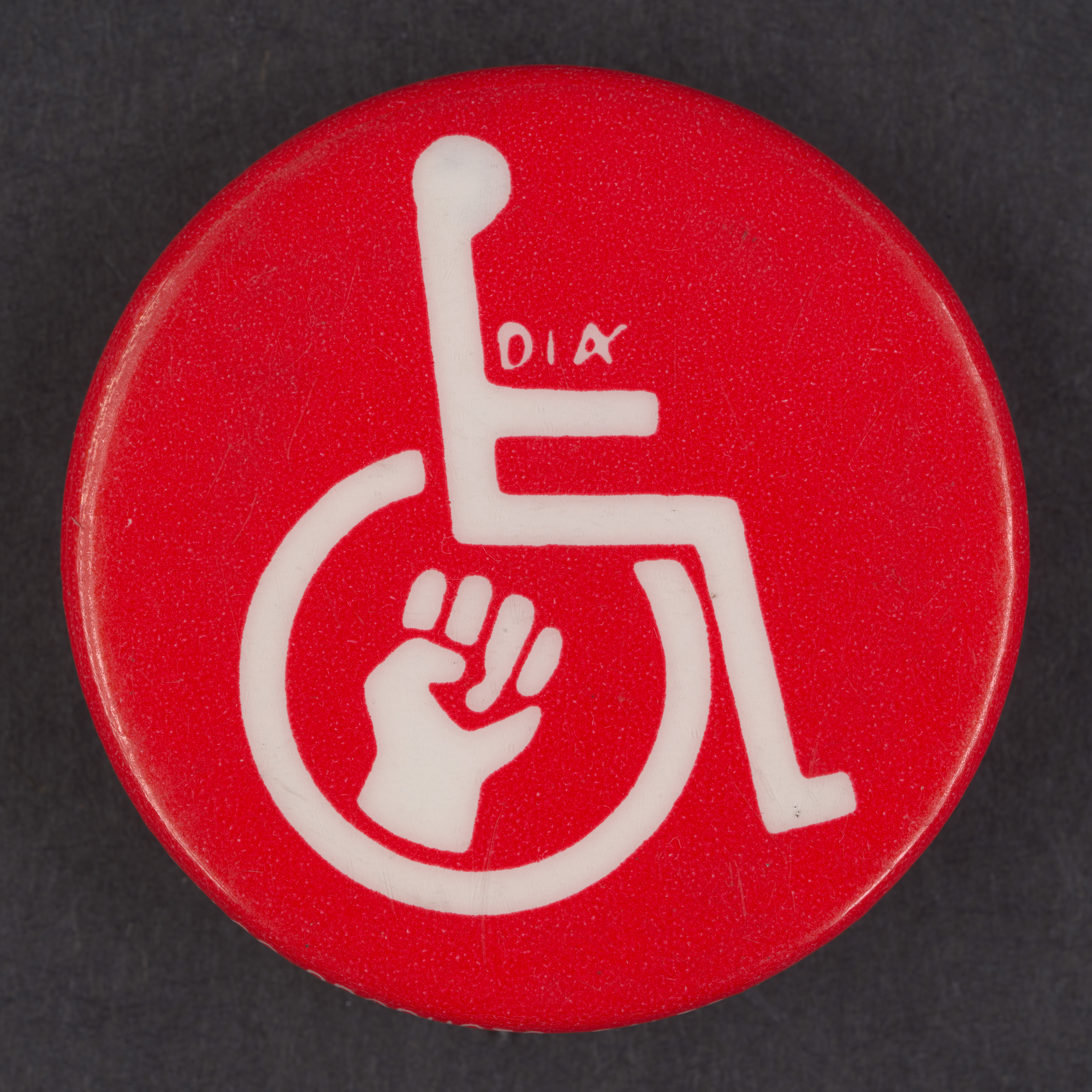 Circular metal button. Red with a white symbol of an outline of a person in a wheelchair. Also in white is a symbol of a fist inside the wheel and the letters "D.A." in small type above the arm.