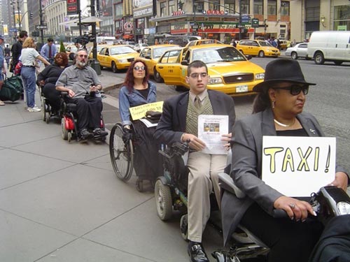 People in wheelchairs are lined up at the edge of a street with signs that say "TAXI." In the street next to them is a row of taxi cabs.