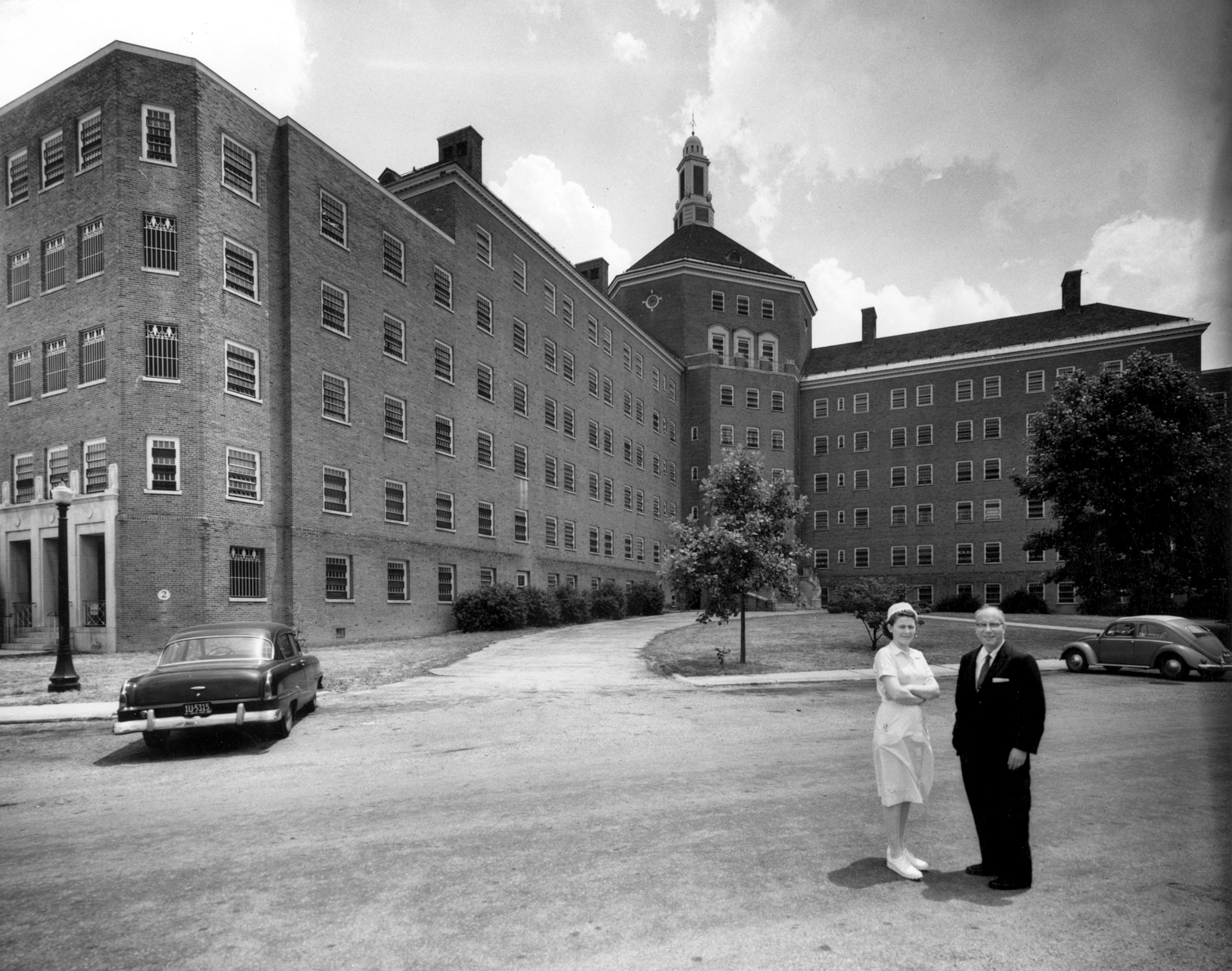 A nurse in a white dress and hat stands next to a man in a dark suit on the far right in the foreground of a black and white photograph. Behind them, taking up the background of the image is a large brick building.