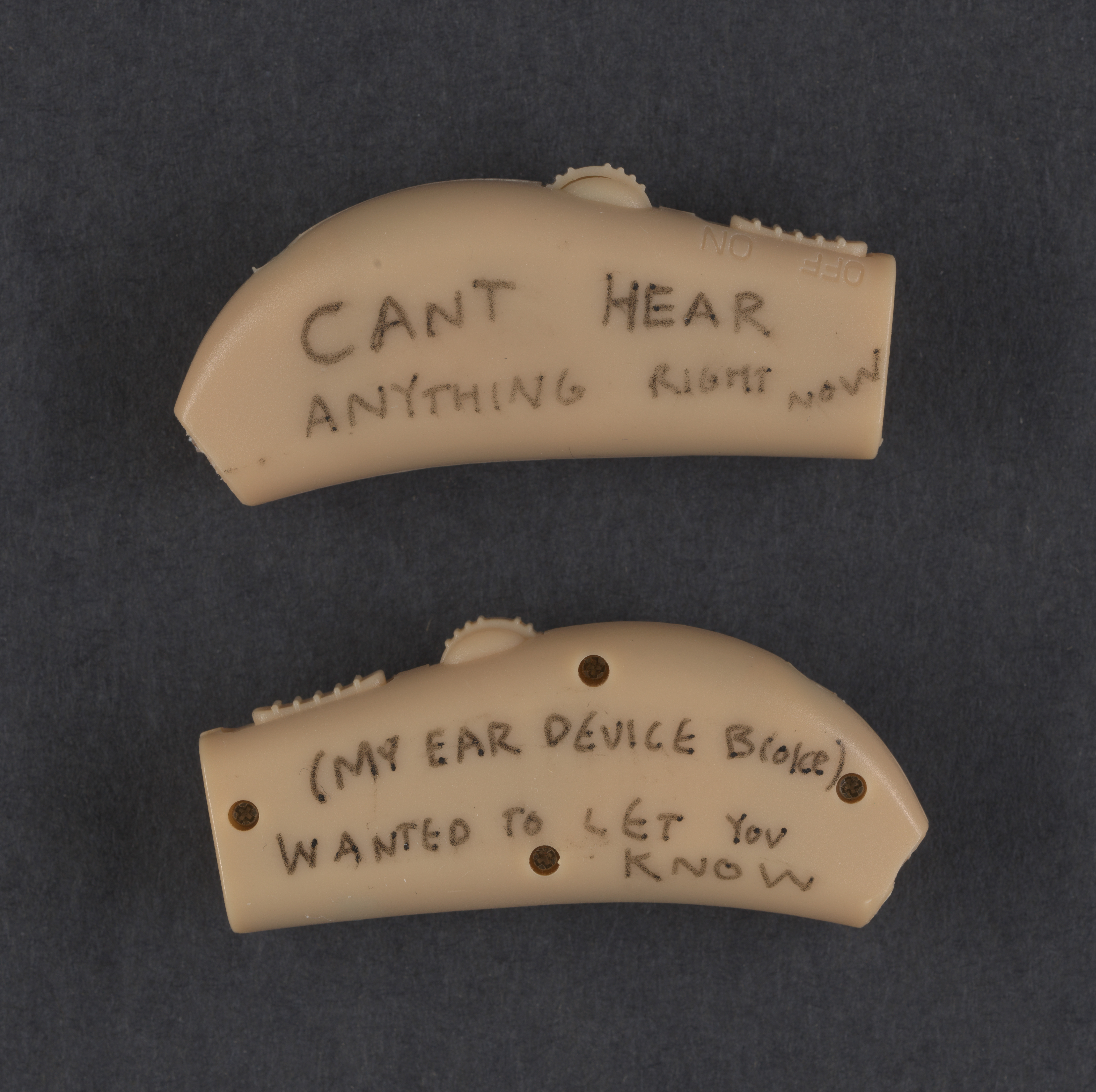 Image of a pair of small beige hearing aids with writing on them. Top reads: "CANT HEAR anything right now" bottom reads: "(MY EAR DEVICE BROKE) wanted to let you know