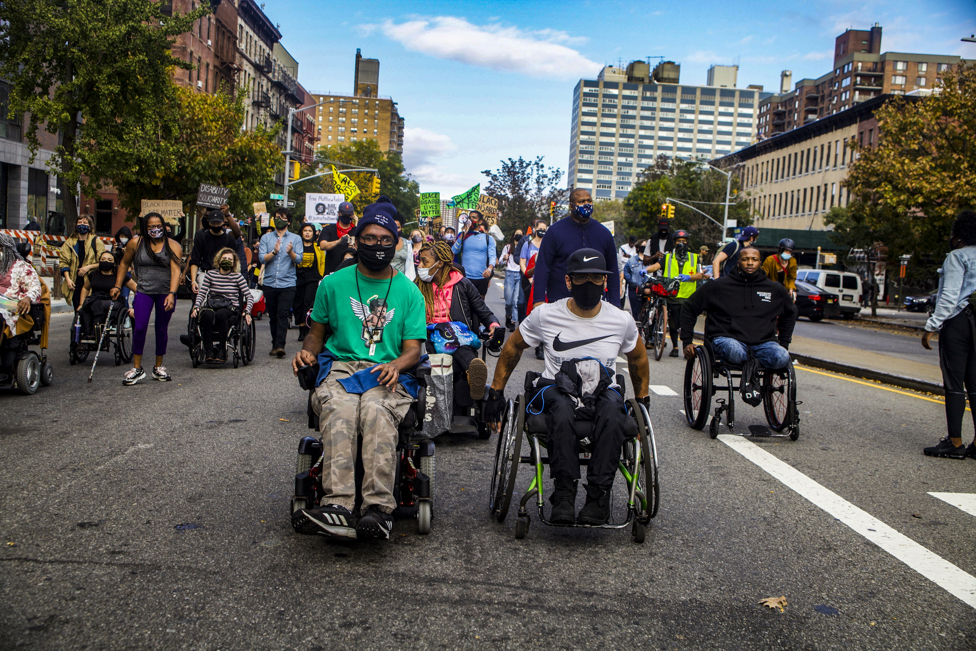 Two men in wheelchairs wearing masks lead a crowd of people, some in wheelchairs or with canes, some holding posters.