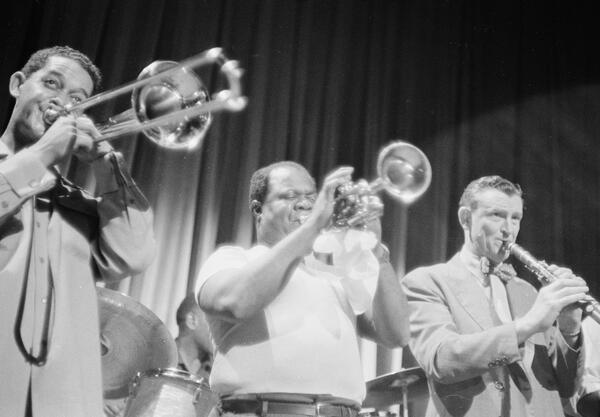 Photograph of Louis Armstrong playing the trumpet on the Jackie Gleason show alongside a clarinet player and a trombone player.