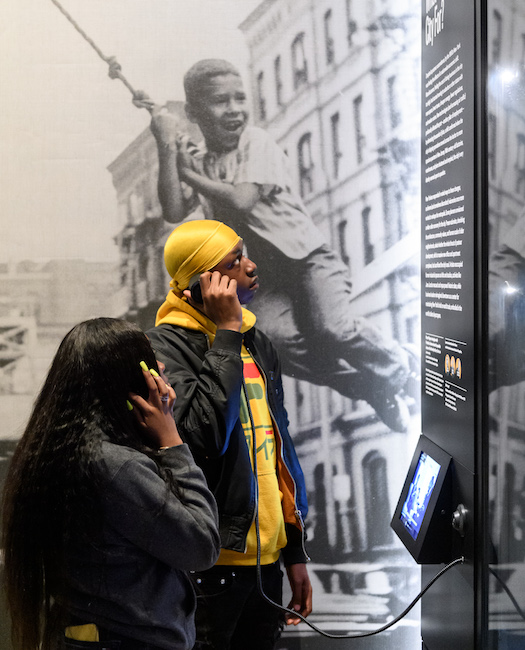 Dois alunos usam fones de ouvido para ouvir uma gravação na galeria "World City", parte de "New York at Its Core".