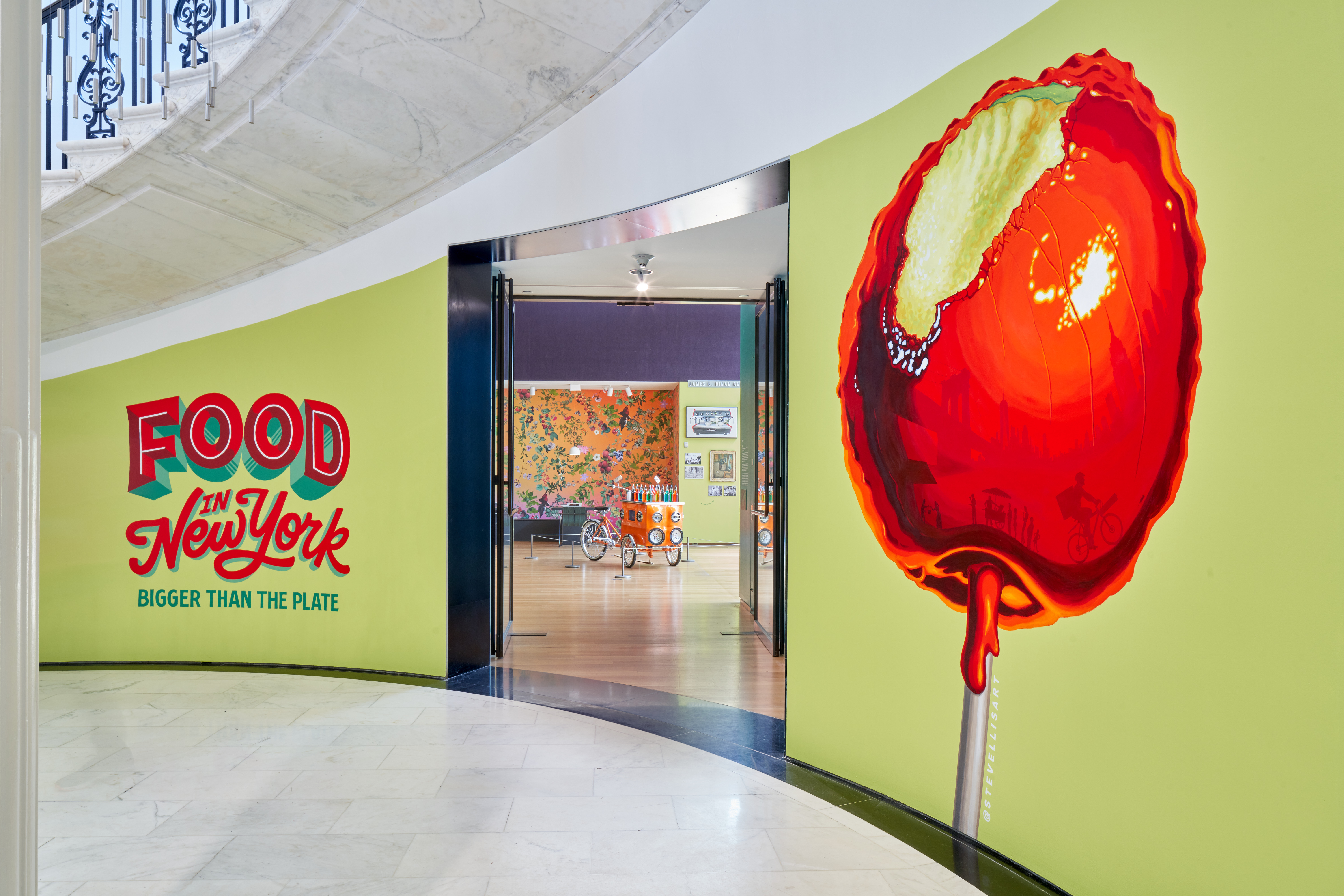 Outside wall of the Food in New York Gallery, a green mural with a red candy apple. Inside the gallery there is a colorful piragua cart.