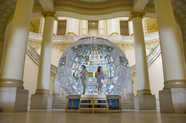 Two women stand in he center of a raised clear sphere, similar to a snow globe, set in a large open room with four white columns.