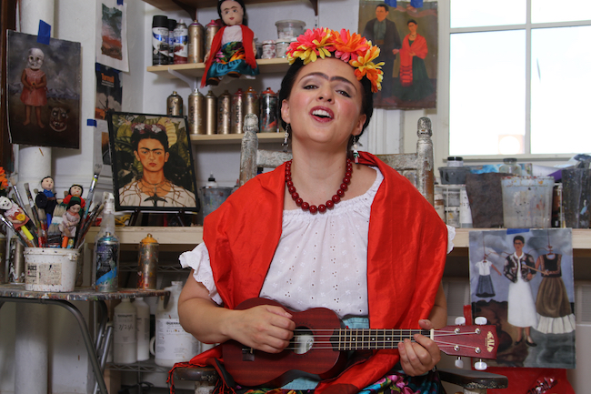 Foto de “The Colors of Frida” do Teatro SEA | Los colores de Frida ”apresentando a encenação da artista de Frida Kahlo em seu estúdio rodeada de pinturas e tocando um instrumento.
