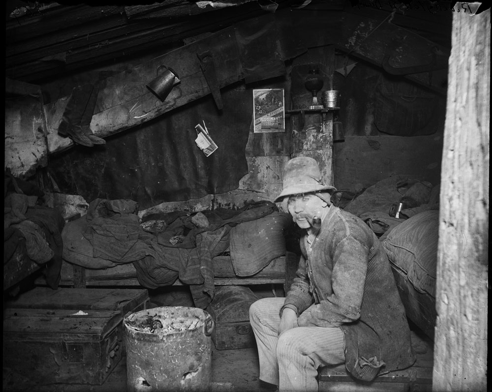 Man sits in a shack-looking house. Both the house and man are dirty, and look uncared for