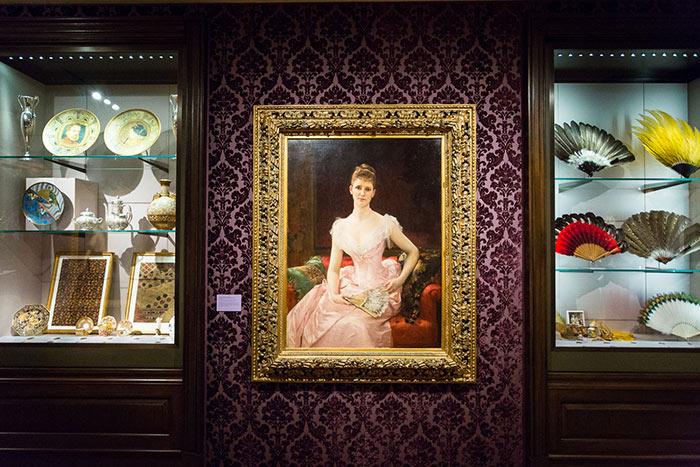 Two exhibition cases containing dishware and fans. In-between them is a painting of a seated woman in a pink dress