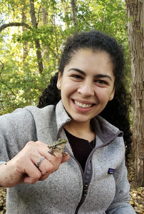 Giselle Herrera가 나무 앞에 서 있습니다. 그녀는 회색 양털 재킷을 입고 있습니다. 그녀는 미소를 지으며 오른손에 개구리를 들고 있습니다.