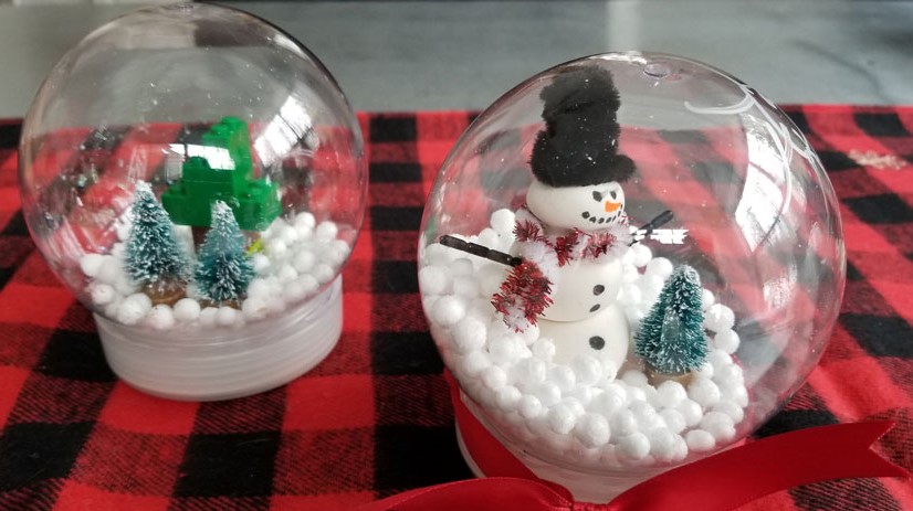 Imagem de dois artesanatos de globo de neve. Uma cena de inverno com árvores sempre verdes à esquerda e um boneco de neve e árvores à direita.