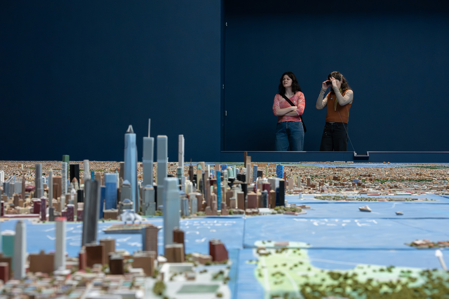 Two people looking at a handmade scale model of New York City through binoculars