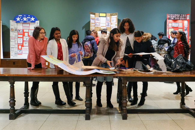 Un groupe d'étudiants se tient sur le côté d'une longue table en bois pour regarder des documents et des projets de la Journée de l'histoire.