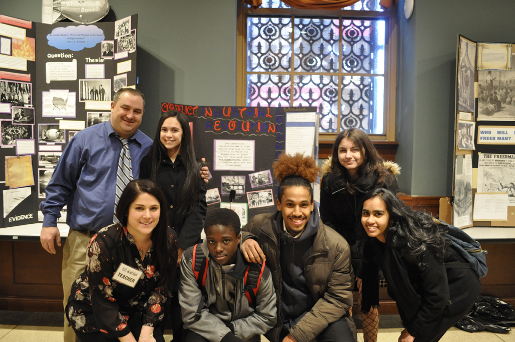 Cinco estudantes do ensino médio e dois professores posam sorrindo para a câmera na frente de um painel de exposições no dia da história da cidade de Nova York em 2018.