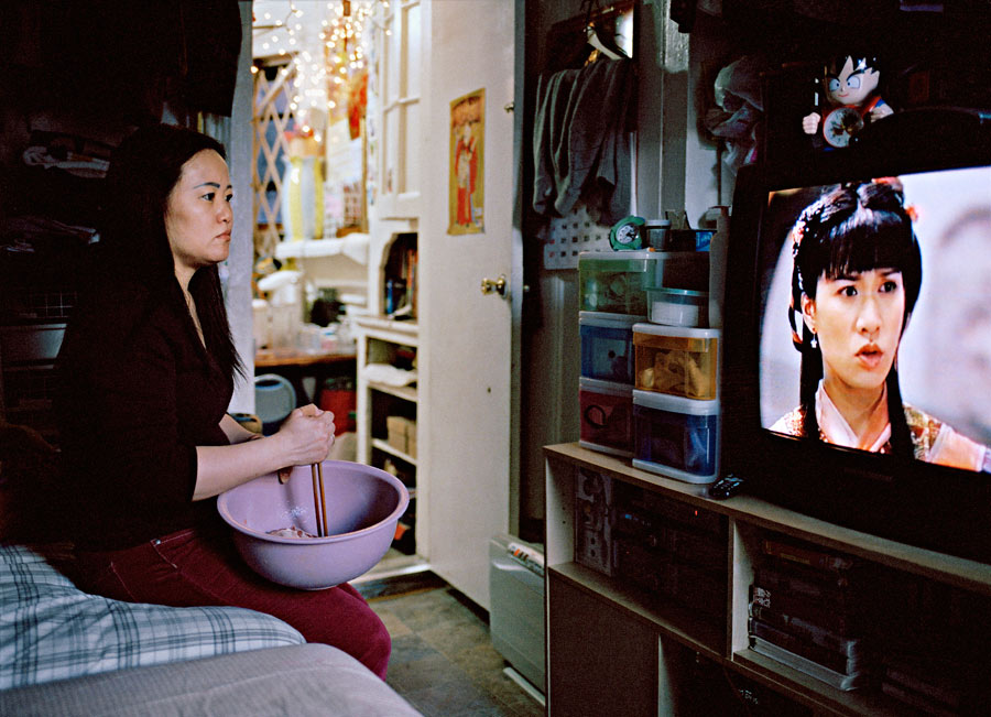 A Chinese woman stirs food in a bowl while watching a Chinese soap opera. She is sitting on a bed in an apartment.