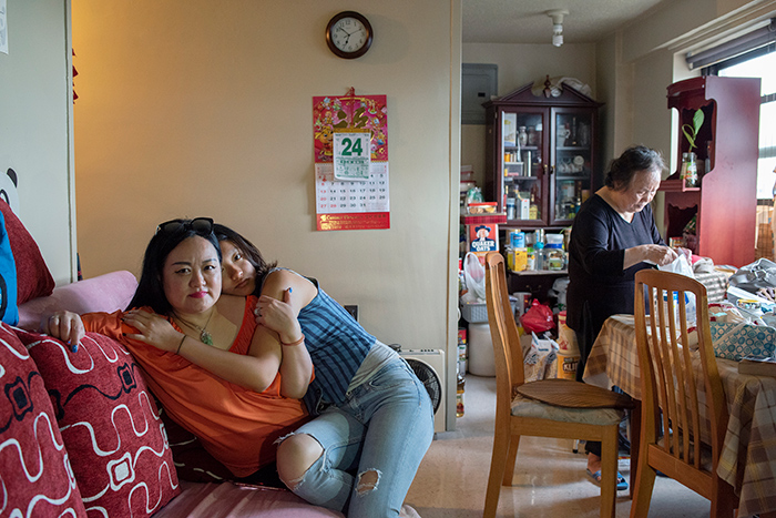 A woman sits on a couch, with her teenage daughter on her lap hugging her. An older woman stands in the background.