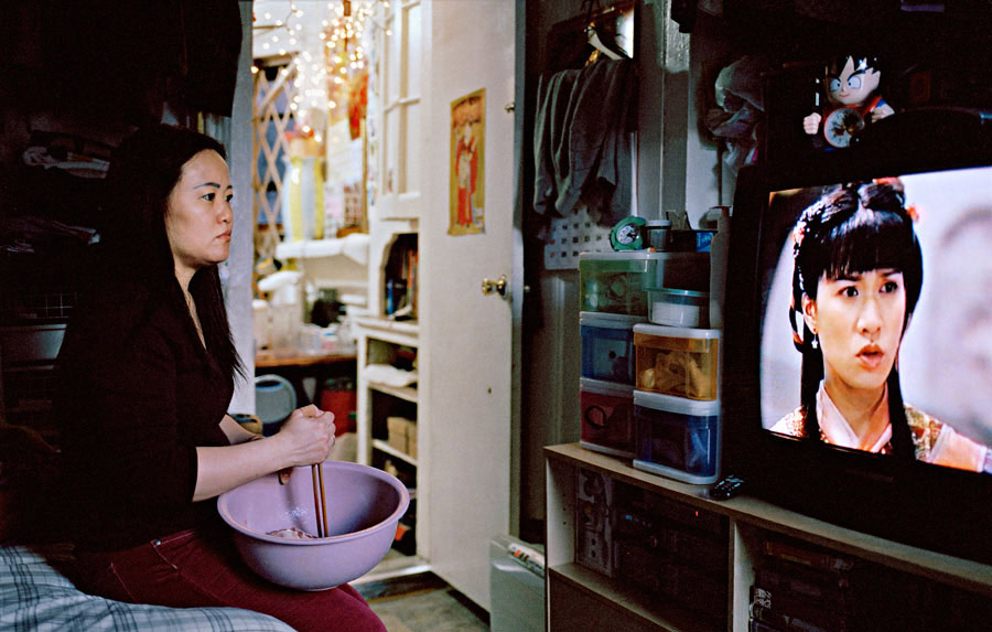 중국 여자는 중국 드라마를 보면서 그릇에 음식을 저어줍니다. 그녀는 아파트에서 침대에 앉아있다.
