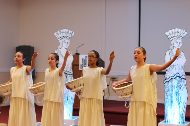 Four girls dressed in Grecian costume stand in front of two handmade caryatids. Mid-performance, they stand with one arm outstretched and the other holding a basket.