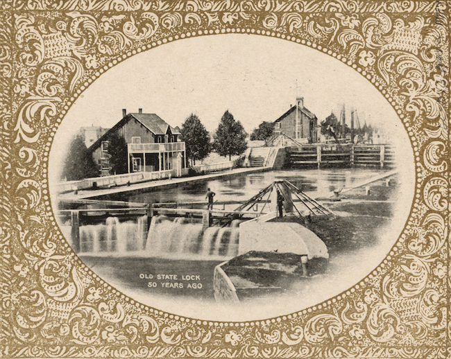Oval-sized image of the Old State Lock-showing buildings and trees near a river with a waterfall in the foreground. Frame is elaborately decorated with a scroll design, brown in color.
