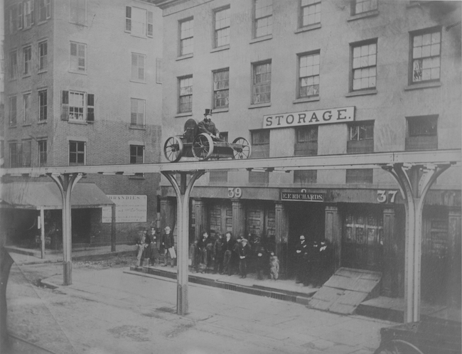 Black and white photo of a man riding in a cart on a elevated platform above a city street.