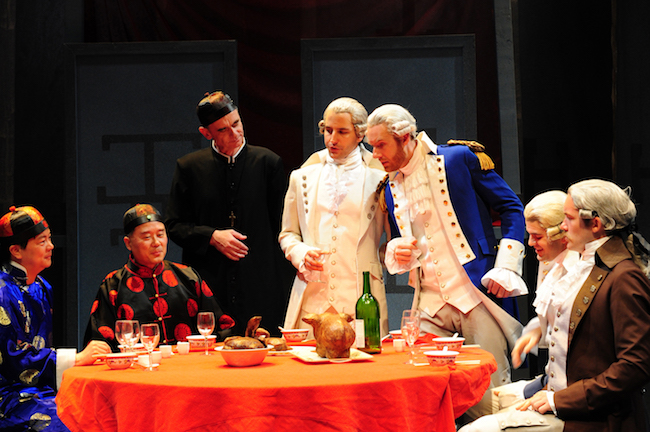 A color photograph of a group of seven costumed actors gathered around a table. Three on the left (two seated and one standing) wear traditional Chinese clothes, while the remaining men (two standing, two sitting) where powdered wigs and colonial dress.