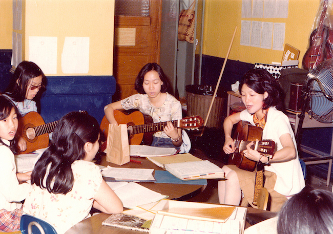 Photograph of a group of women sitting around a table in a classroom-like setting, all holding guitars with papers in front of them.