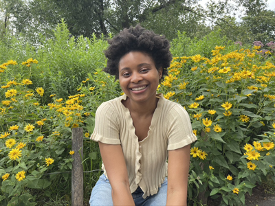 Kadi Ba está sentada e sorrindo em frente a um arbusto de flores amarelas. Ela está vestindo um cardigã de mangas curtas bege claro e jeans azul claro.