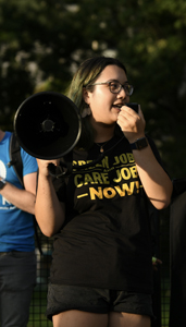 Kathryn Gioiosa is standing holding a megaphone. She is wearing a black t-shirt that reads, “Green Jobs! Care Jobs! Now!” and black shorts. 