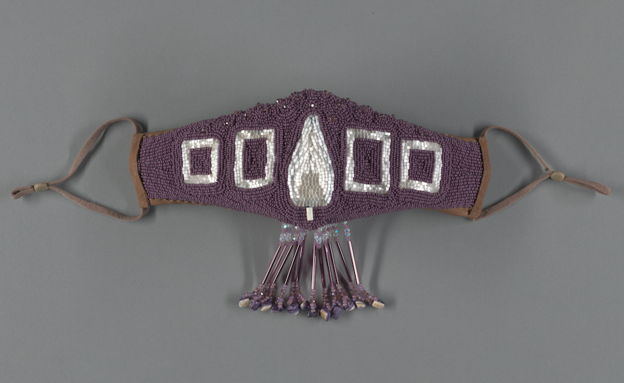 A face mask made with a beaded purple background and white beaded design. A small fringe hangs down from the center.