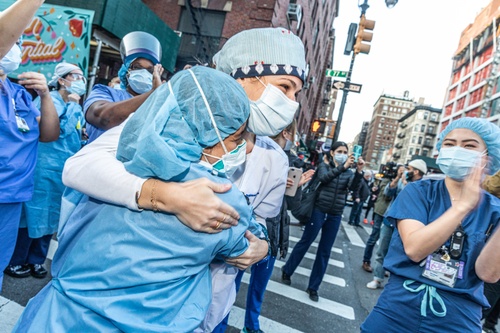 Color photograph of two women in full scrubs and protective gear hugging on a NYC street with other individuals, also in scrubs, surrounding them.