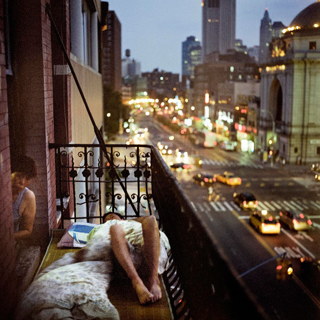 A person lies on the terrace of an apartment building, while a man leans out a window to look at the camera