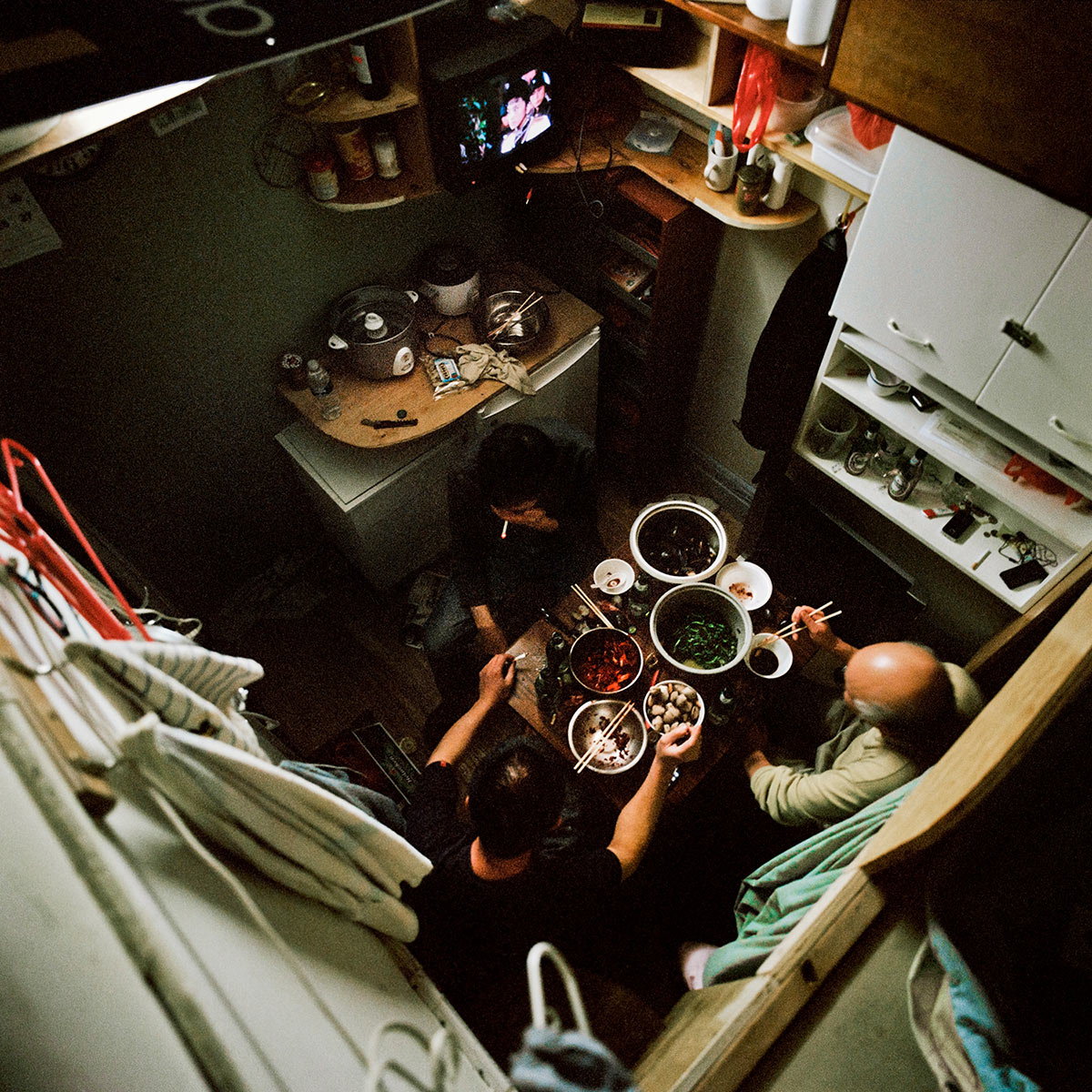 Three men sit around a low table in a makeshift kitchen. There are multiple bowls on the table and the men are eating.