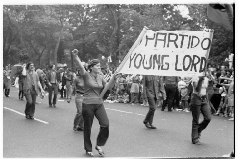 People march during a parade, while two hold a sign between them that says “_PARTIDO/YOUNG LORD”