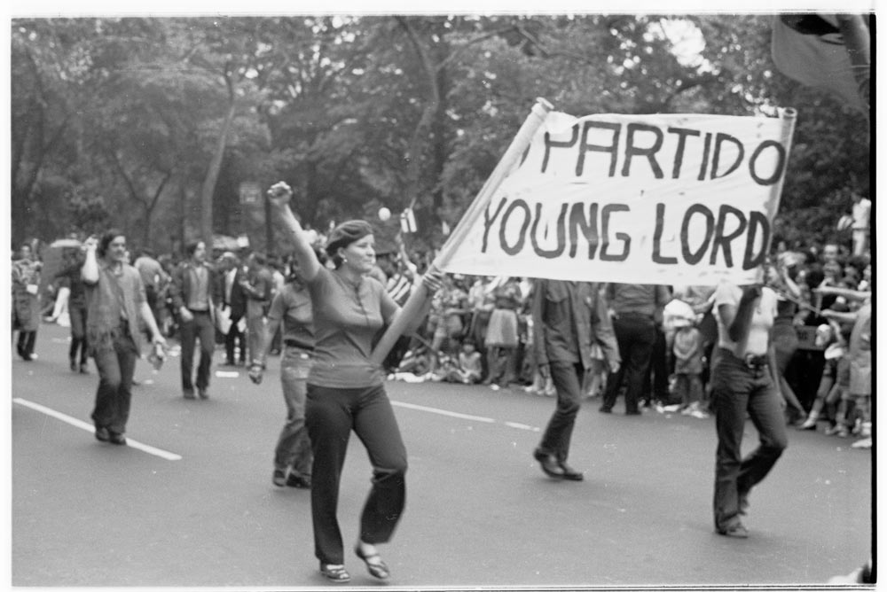 La gente marcha durante un desfile, mientras dos sostienen un cartel entre ellos que dice "_PARTIDO / YOUNG LORD"