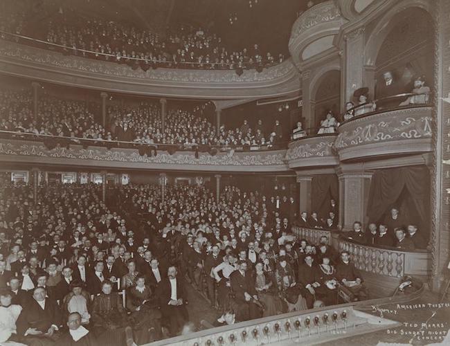 Photograph of a theater taken from the stage looking out at a full audience, showing the orchestra, mezzanine, balcony, and box seats on the right hand side.