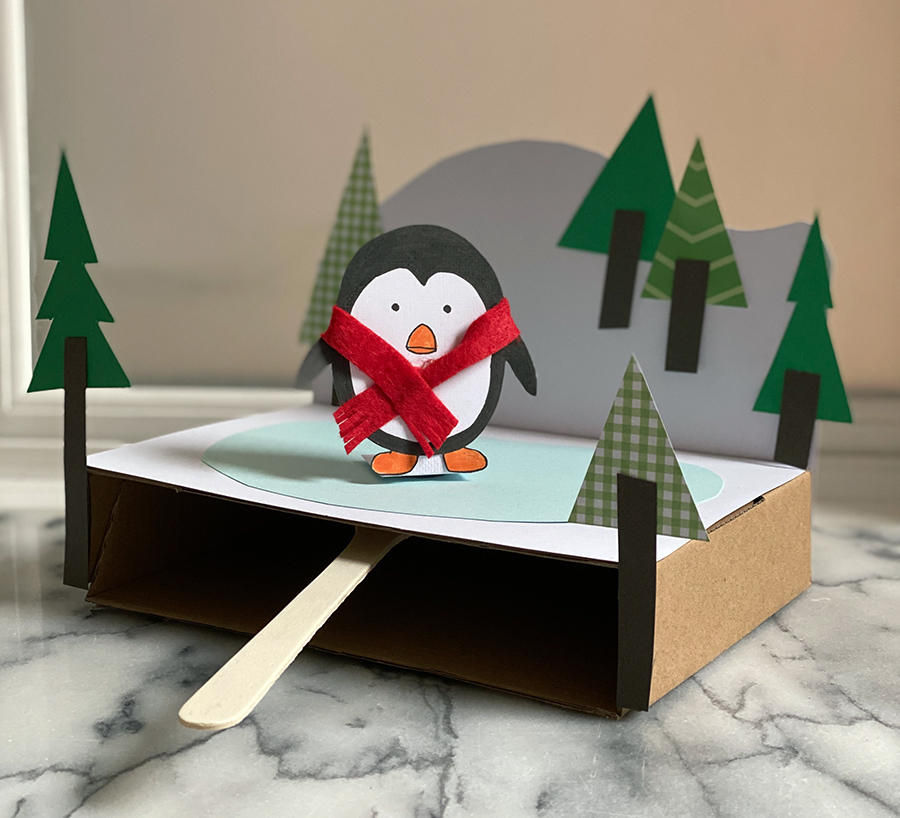 A photo of a craft project using paper and cardboard to create a penguin wearing a scarf skating on a frozen field of ice. 