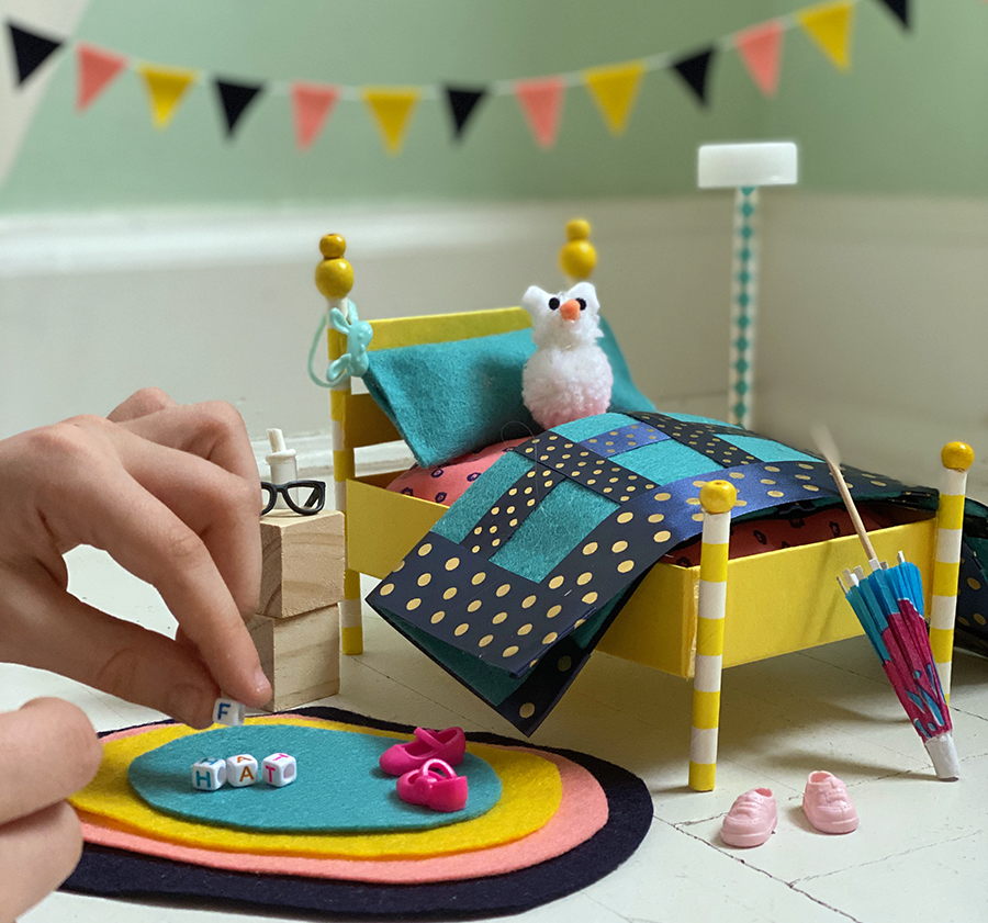 A photograph of a colorful set of miniature bedroom furniture, including a bed with stuffed animal, nightstand, rug, and decorations. An adult’s hand holding a tiny block is entering the view from the left, as if about to place it on the rug.