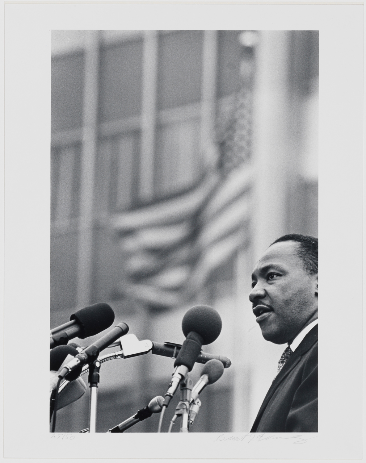 Fotografia em preto e branco de Martin Luthur King Jr. em pé ao microfone com um prédio e uma bandeira balançando atrás dele.