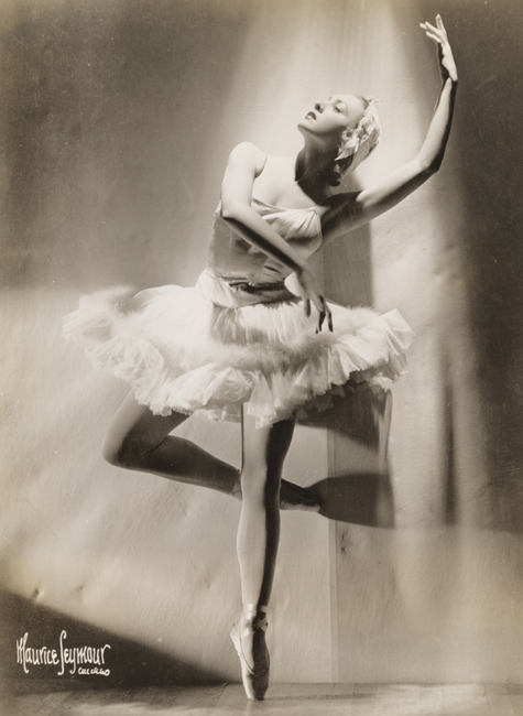 A ballerina, in costume, stands en pointe with one leg half bent at the knee and her arms raised.