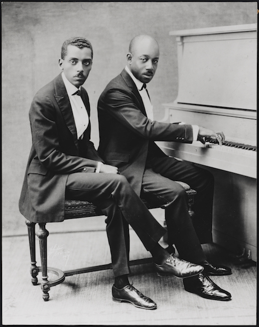 Deux messieurs afro-américains en costume sont assis sur un banc de piano, avec un piano à droite. L'homme de droite a les mains sur le clavier.