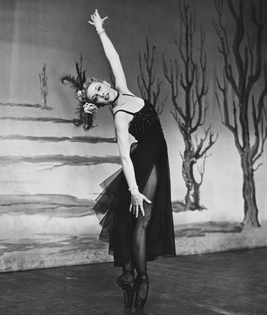 A ballerina, in costume, stands en pointe with one arm raised straight above her head, in front of a set backdrop with a line of barren trees.