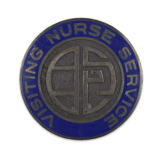 Metal lapel pin. An abstract design in plain metal in the center is surrounding by a blue enamel ring with the words "Visiting Nurse Services"